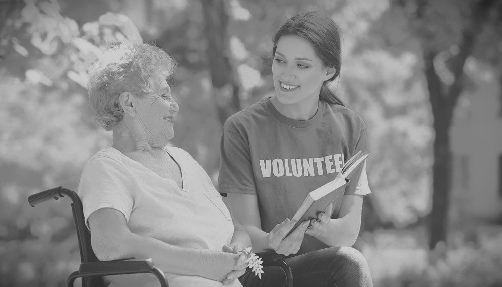 Young Female Volunteer Reading Book To Senior Woman On Wheelchair Outdoors